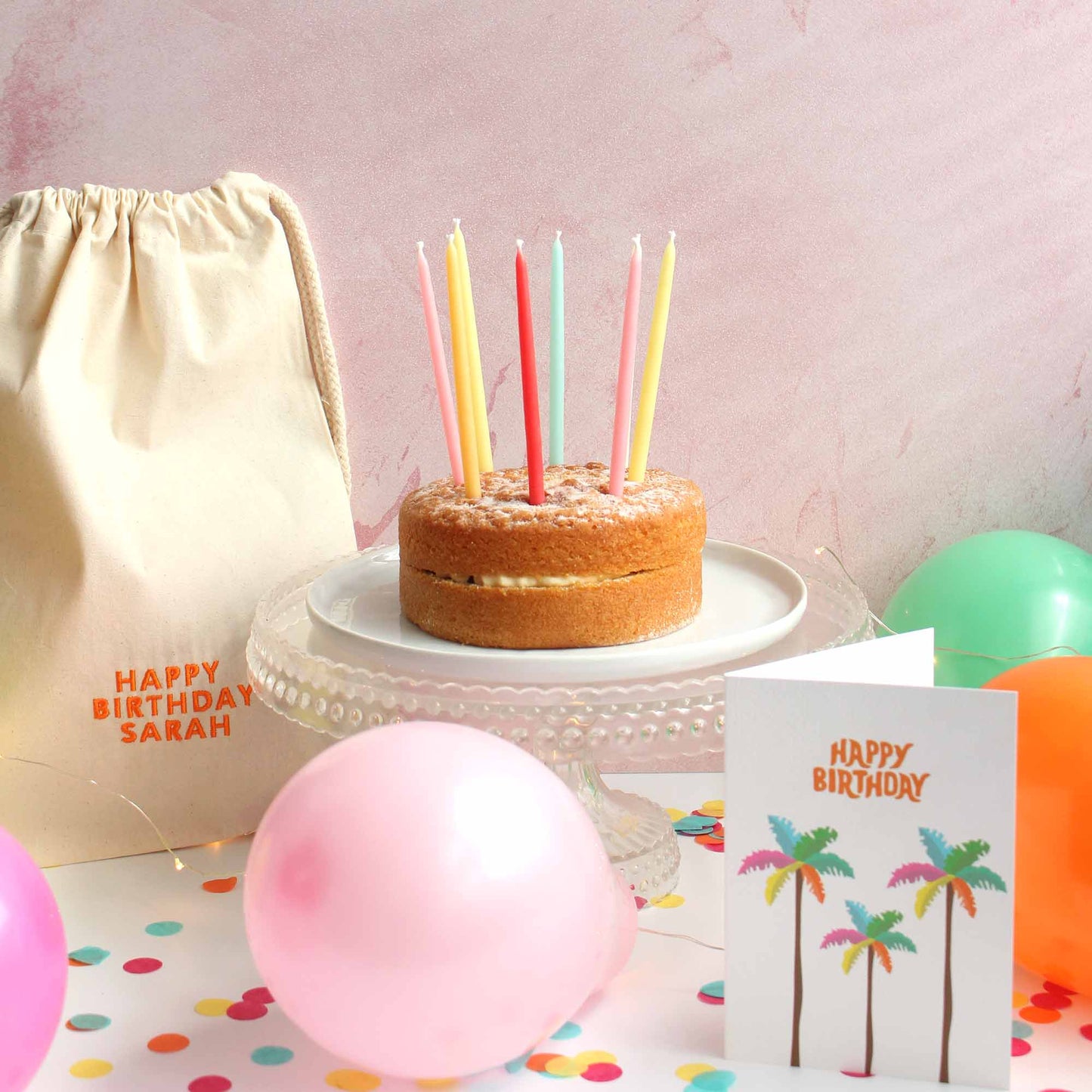 Image of birthday special scene with a cake with colourful beeswax candles, a personalised drawstring bag, colourful balloons, greeting cards and confetti all on a white table in front of a pink wall.