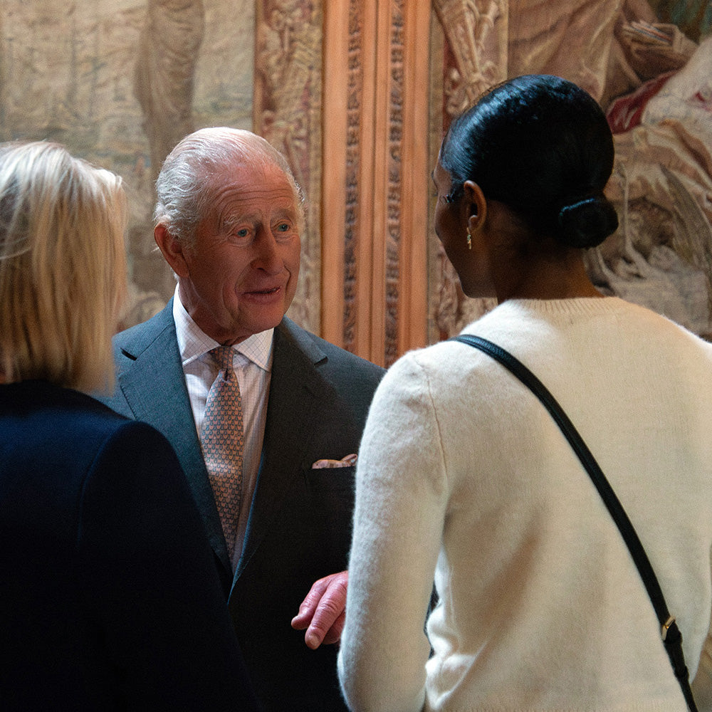 King Charles in a dark suit and patterned tie conversing with Tihara Smith in an indoor setting with a large classical tapestry in the background.