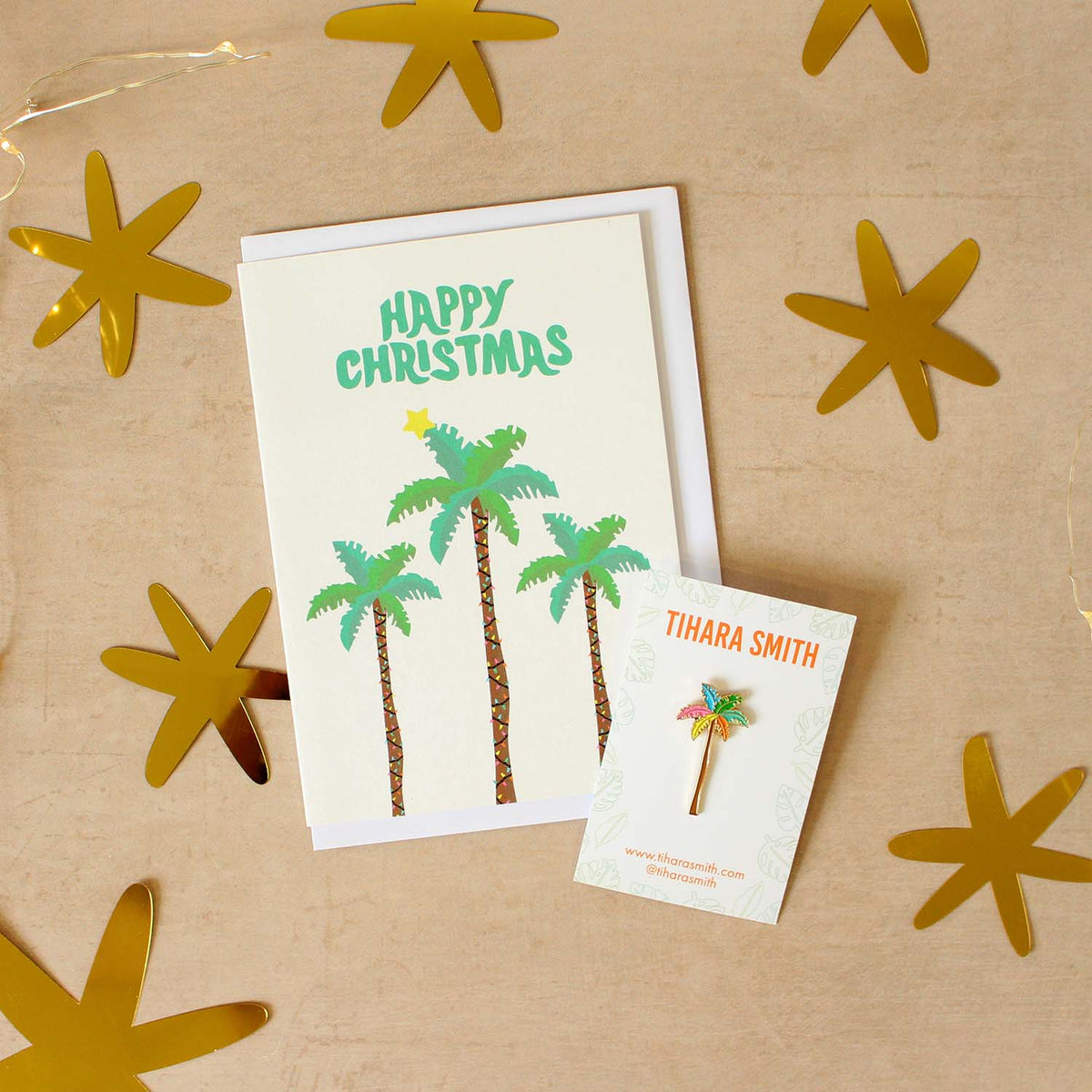 Image of a palm tree Christmas card and palm tree pin on a textured beige surface with star sequins around it.