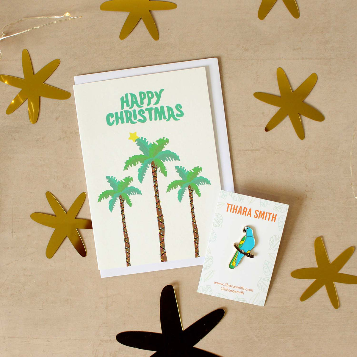 A palm tree Christmas card and parrot pin on a beige surface surrounded by cut out gold stars.