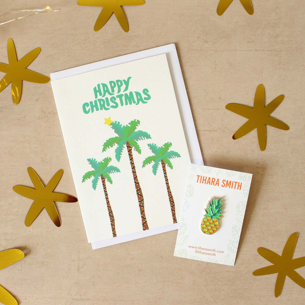 A palm tree Christmas card and pineapple pin on a beige surface surrounded by cut out gold stars.