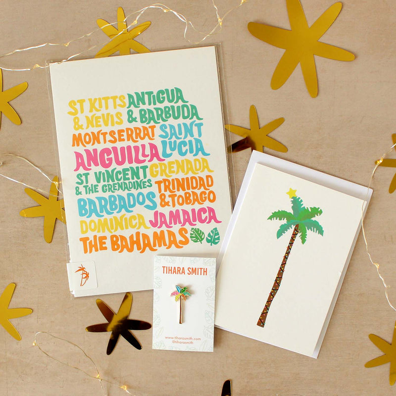 A styled flat-lay of tropical-themed stationery on a beige background with star cutouts and fairy lights. The set includes a Caribbean islands print, a Christmas palm tree greeting card, a palm tree pin, and an envelope.