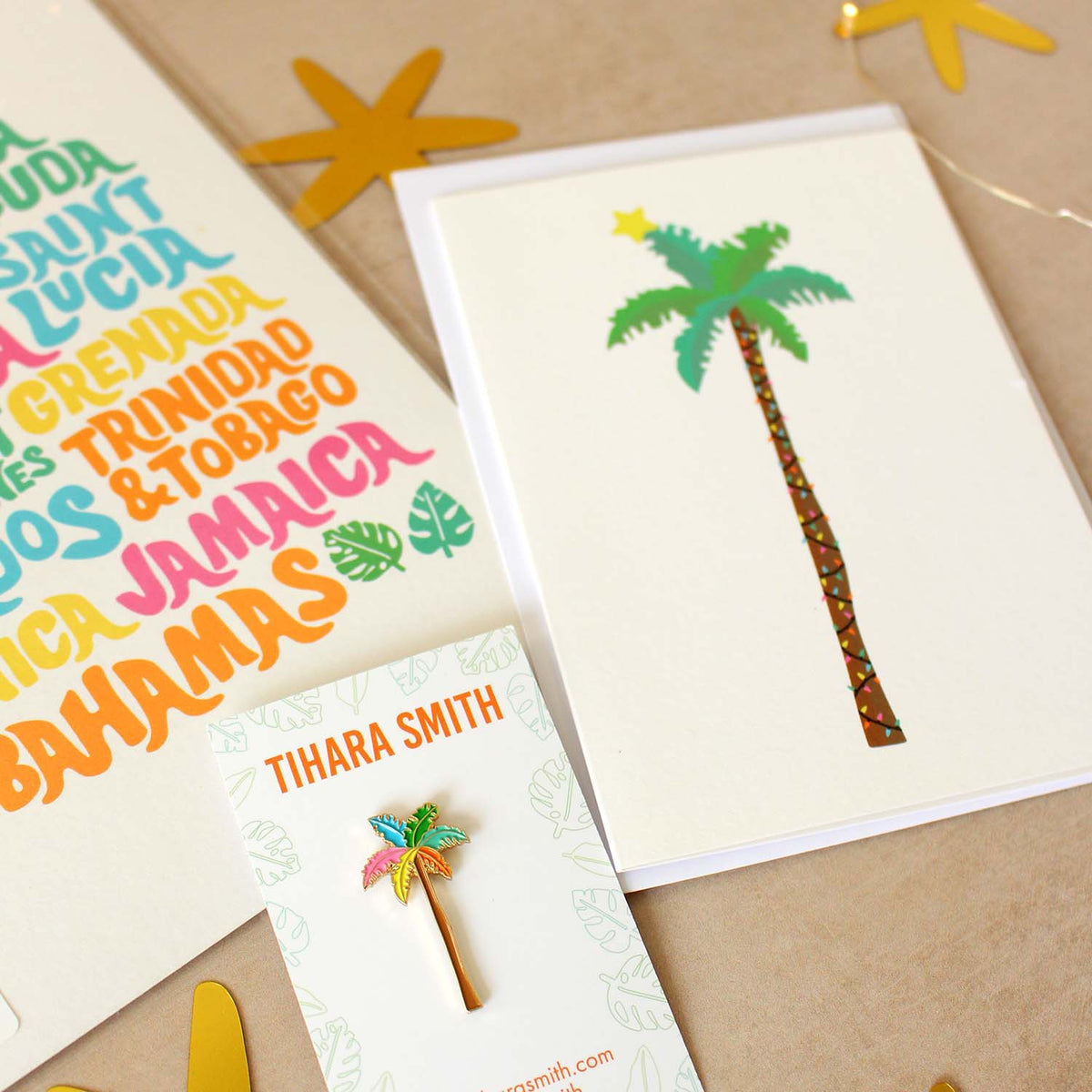 A styled flat-lay of tropical-themed stationery on a beige background with star cutouts and fairy lights. The set includes a Caribbean islands print, a Christmas palm tree greeting card, a palm tree pin, and an envelope.