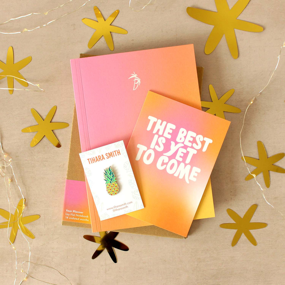 A styled flat-lay of a pink and orange gradient notebook, a matching card that reads 'THE BEST IS YET TO COME', and a  pineapple pin, arranged on a beige surface with gold star cutouts and string lights.