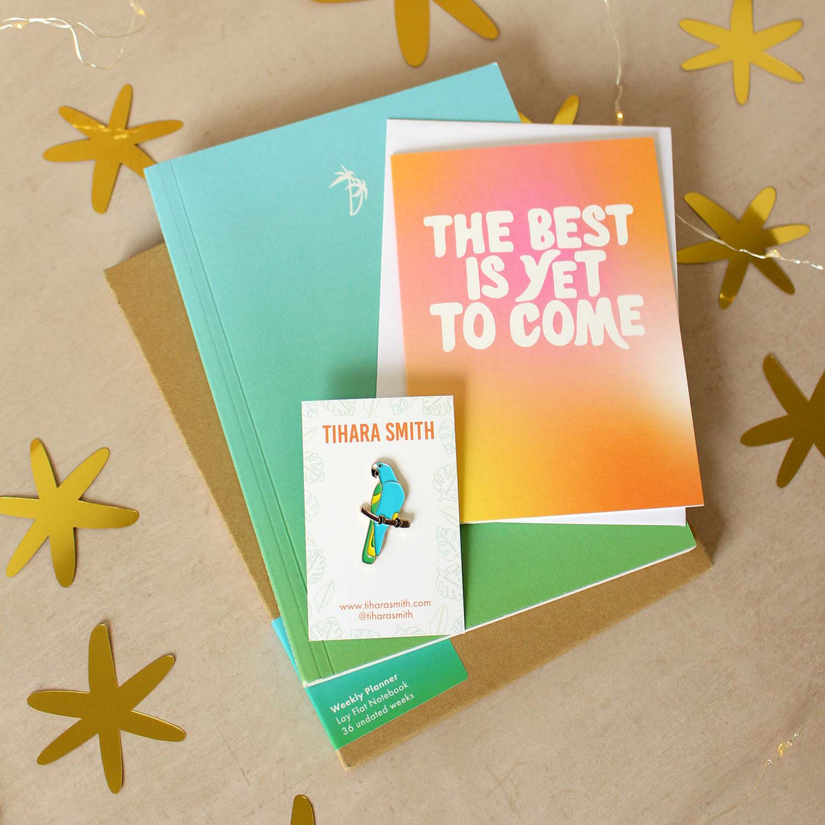 A styled flat-lay of a green and blue gradient notebook, a matching card that reads 'THE BEST IS YET TO COME', and a  parrot pin, arranged on a beige surface with gold star cutouts and string lights.