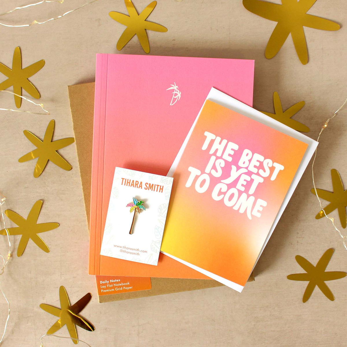 A styled flat-lay of stationery and a small enamel pin on a beige background with gold star cutouts and string lights. The set includes a pink to orange gradient notebook, a greeting card reading 'THE BEST IS YET TO COME,' a palm tree pin.