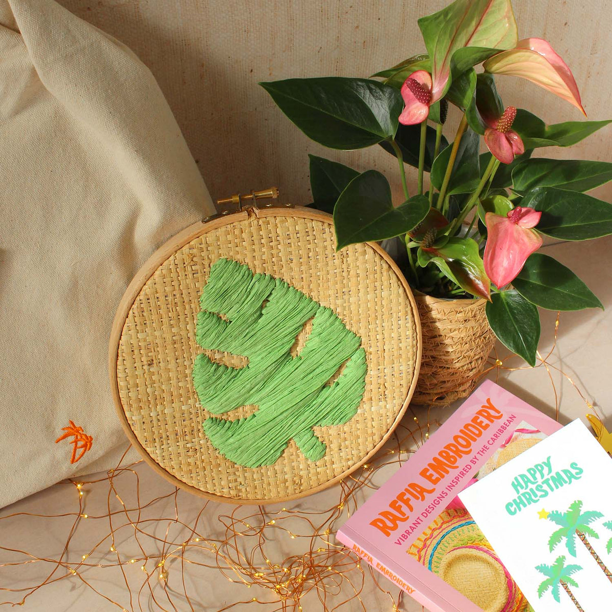 A round wooden embroidery hoop containing a green monstera leaf design stitched on raffia fabric, styled with a potted anthurium plant, a cream canvas bag, fairy lights, and the Raffia Embroidery book.