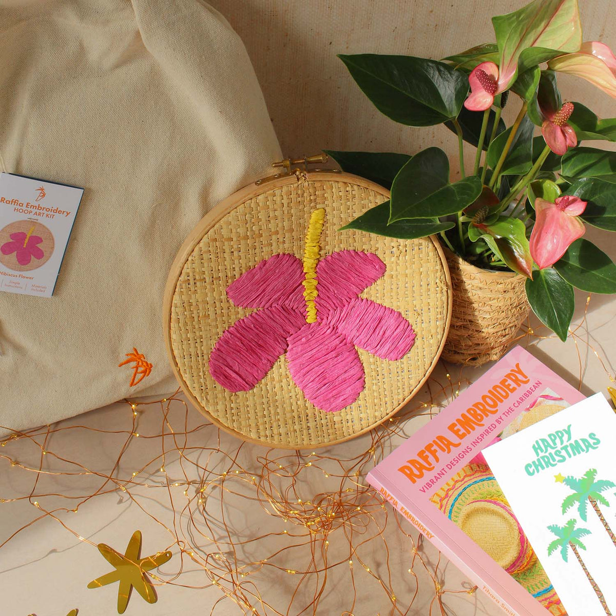 A round embroidery hoop with a pink hibiscus flower raffia embroidery design and a yellow stem, styled on a beige surface with a canvas bag, plant, star decor, and printed card.