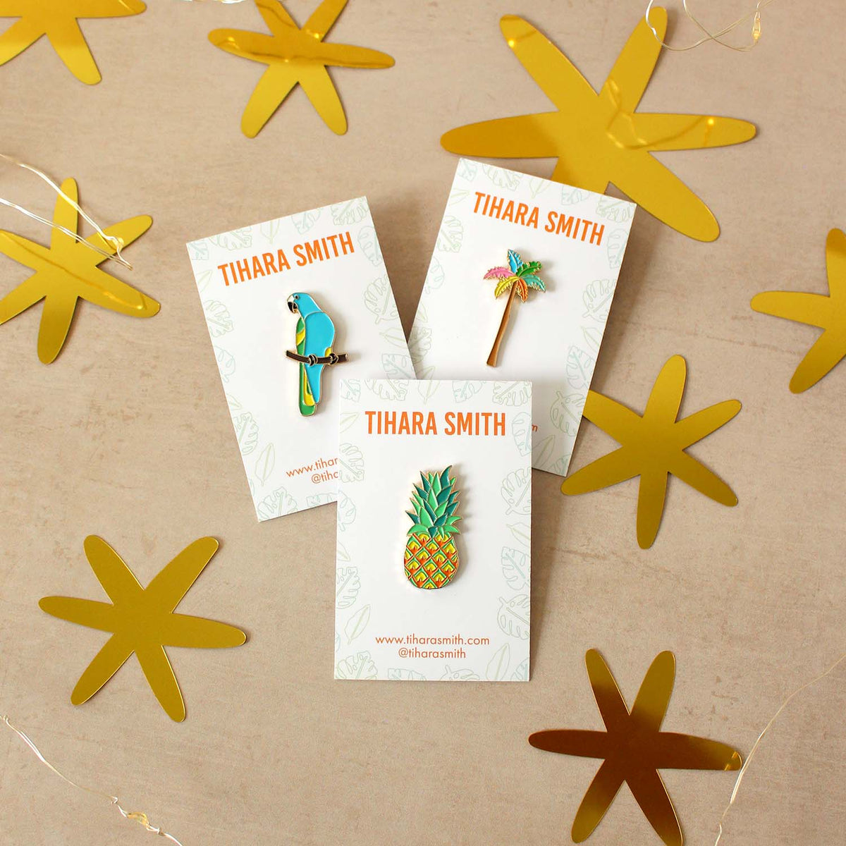 A parrot pin, pineapple pin and palm tree pin on a beige surface with gold star confetti.
