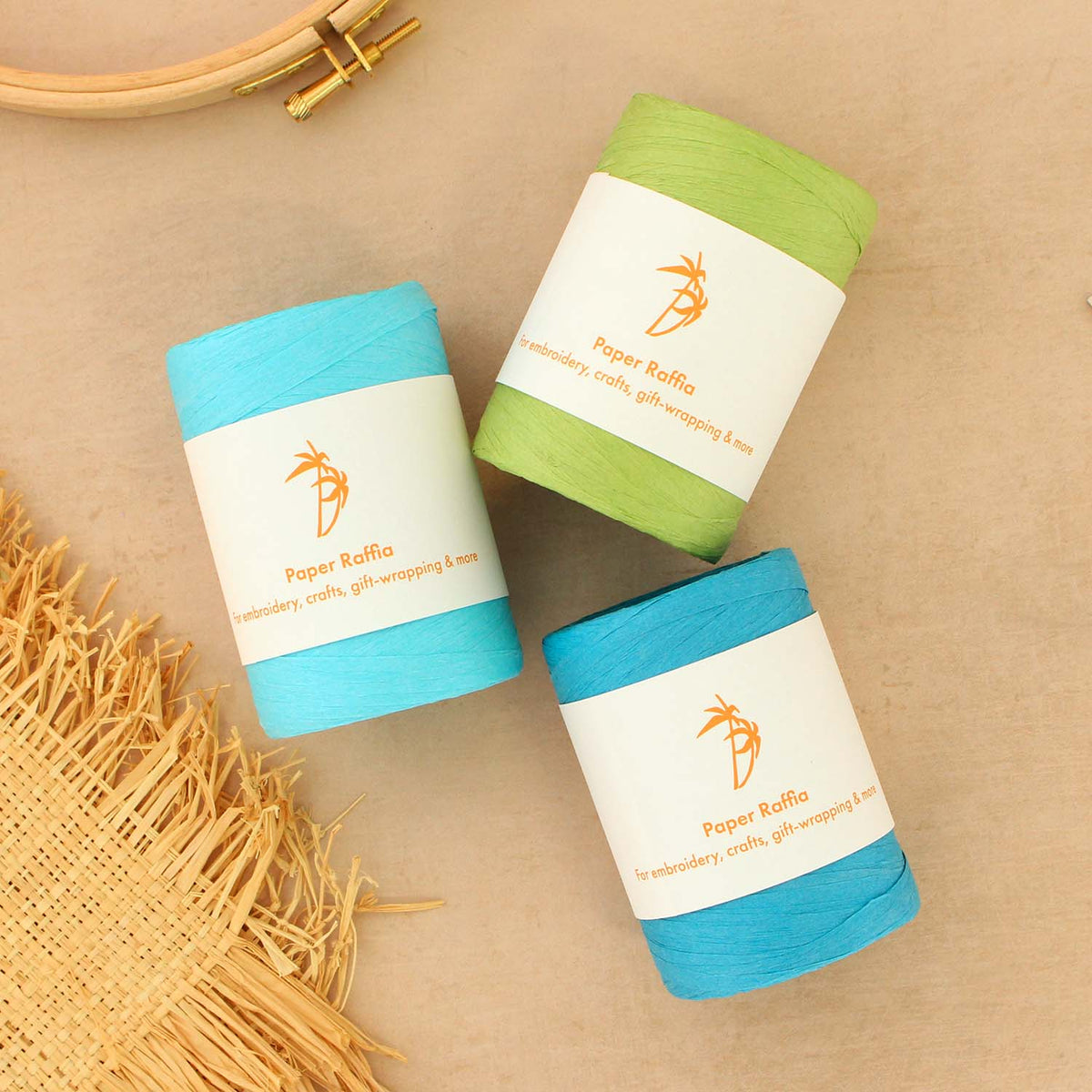 Three spools of Paper Raffia embroidery yarn in light blue, scallion green, and blue, arranged on a beige textured background with decorative raffia fabric and an embroidery hoop.