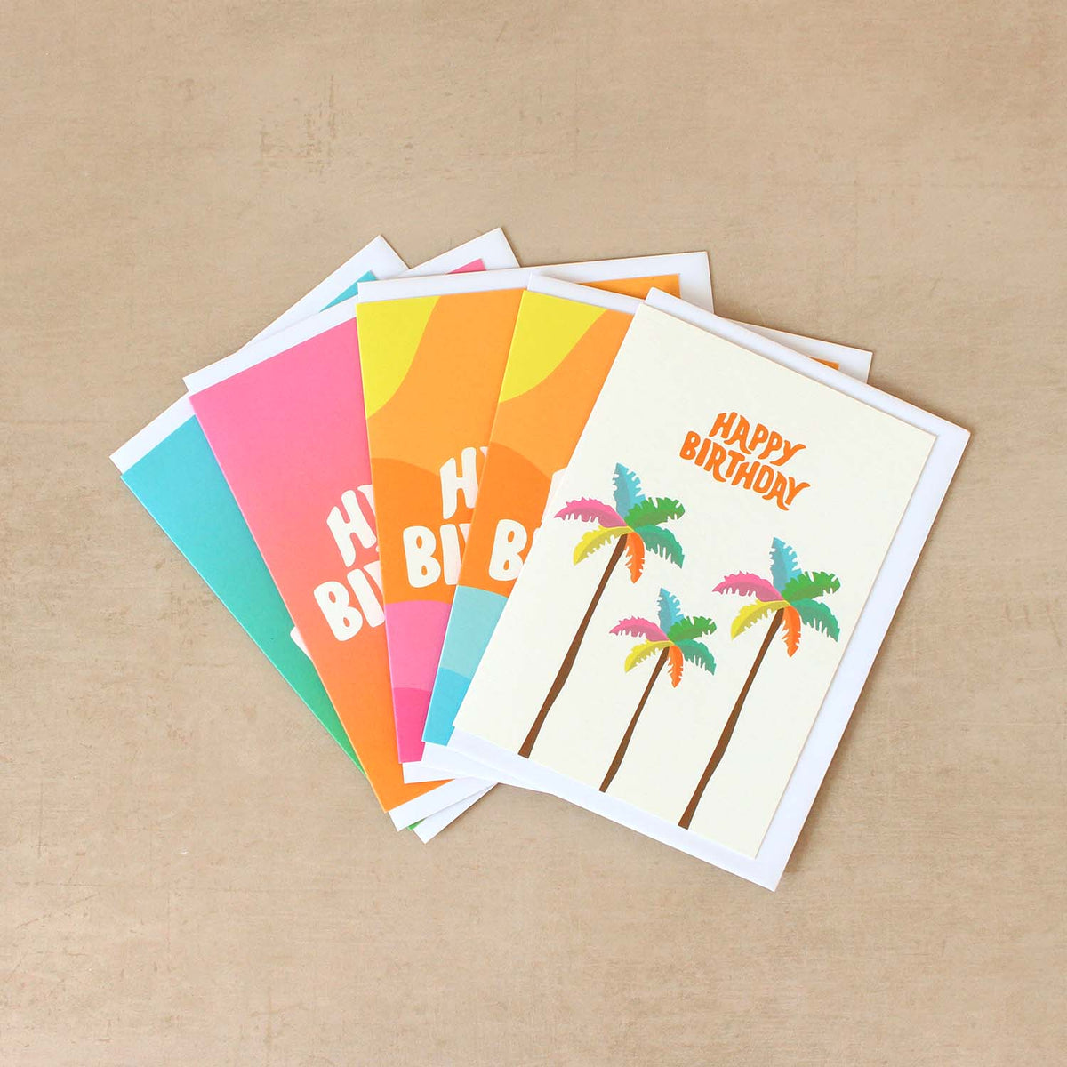 A fan of colourful birthday greeting cards arranged on a light beige textured surface. The front card is off-white with orange 'HAPPY BIRTHDAY' text and three palm trees in pink, blue, yellow and green leaves.