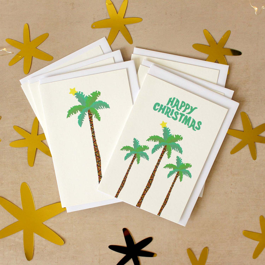Stack of six palm tree Christmas cards on a beige surface with star cut outs around them.