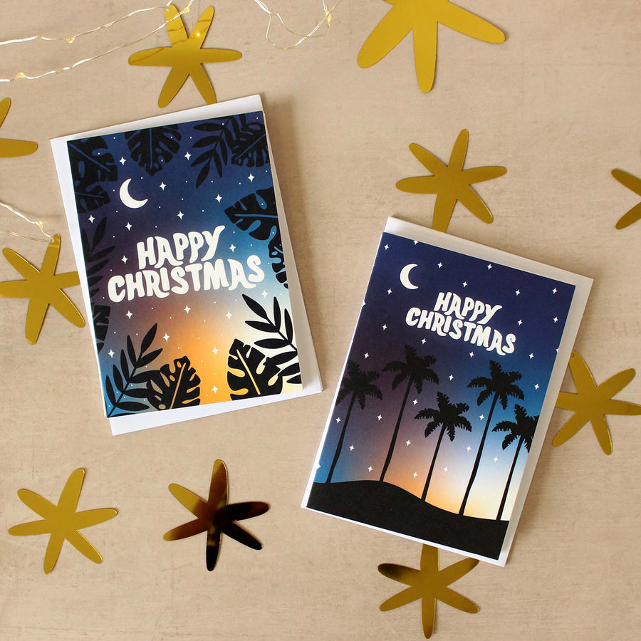 Stacks of tropical Christmas cards on a beige surface with gold cut out stars around them. 
