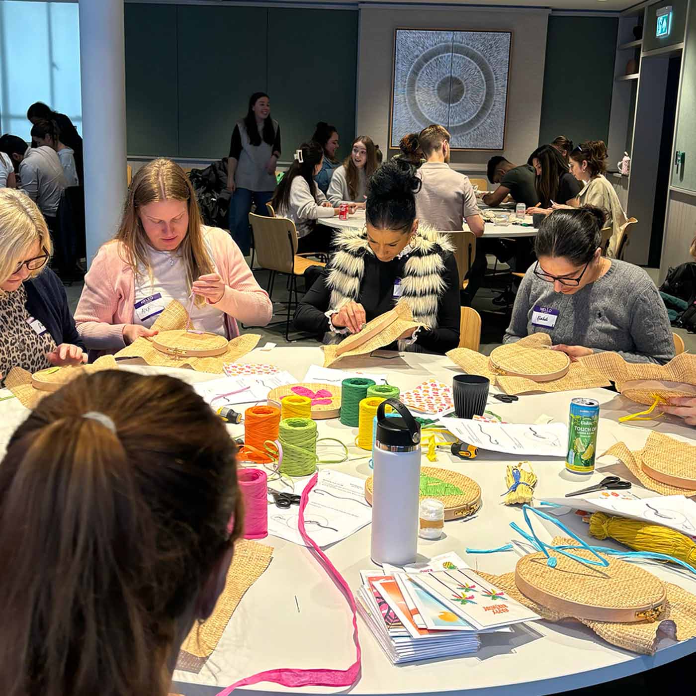 A group of people seated around a table in a raffia embroidery corporate wellbeing workshop, crafting raffia embroidery hoops with colourful raffia thread, papers, and tools on a white table surface.
