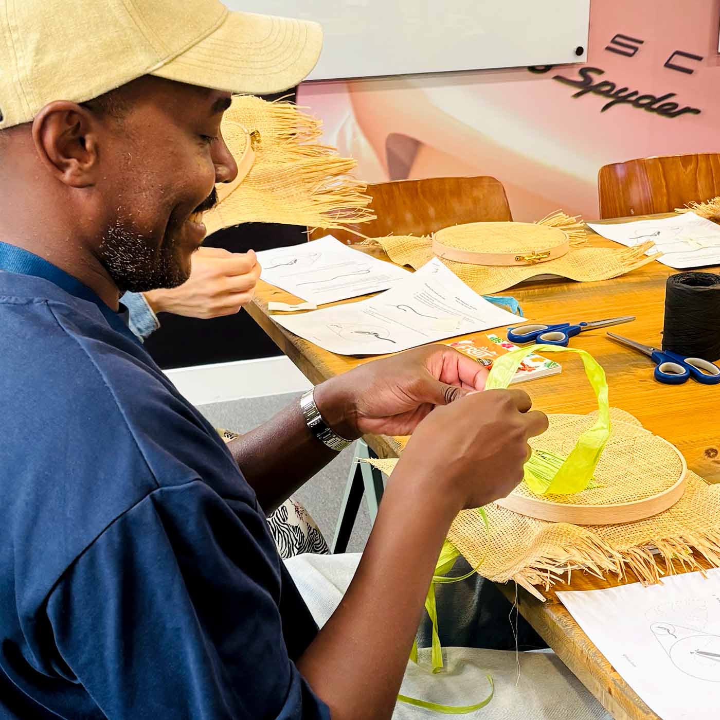 A man wearing a beige cap and navy blue shirt sits at a wooden table at a corporate wellbeing workshop, actively crafting a raffia embroidery hoop with a bright yellow-green ribbon. The table is cluttered with embroidery hoops, scissors, thread, and paper templates.