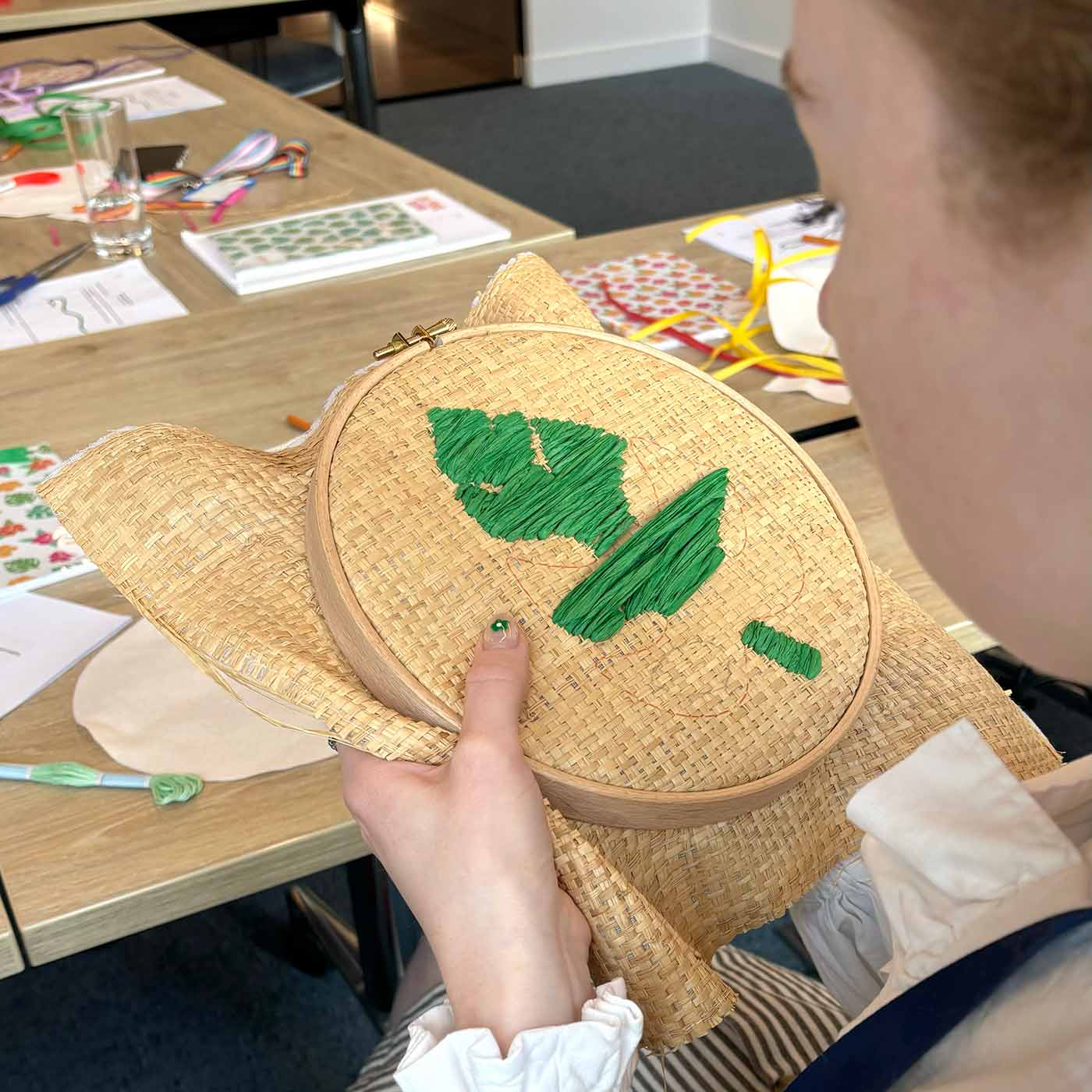 A person holding a raffia embroidery hoop at a corporate wellbeing workshop table, with green raffia stitched on the raffia.