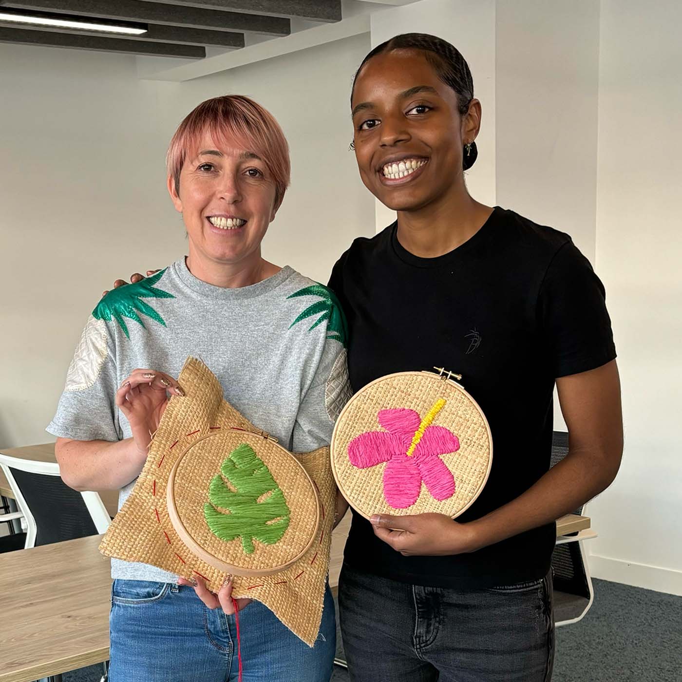 Two smiling women standing indoors at a corporate wellbeing workshop and holding round raffia embroidery hoops. 