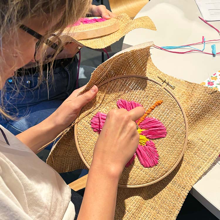 A close-up of a person's hands and head at a corporate wellbeing workshop as they embroider a bright pink flower with yellow and orange details inside a raffia embroidery hoop at a white table.