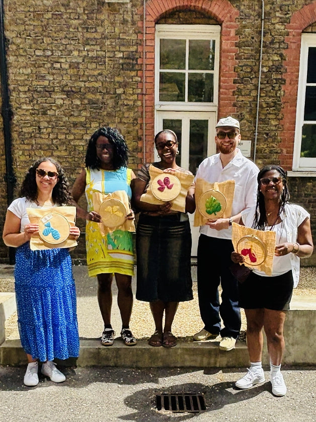 Five people standing on steps in front of a brick building, holding raffia embroidery hoops.