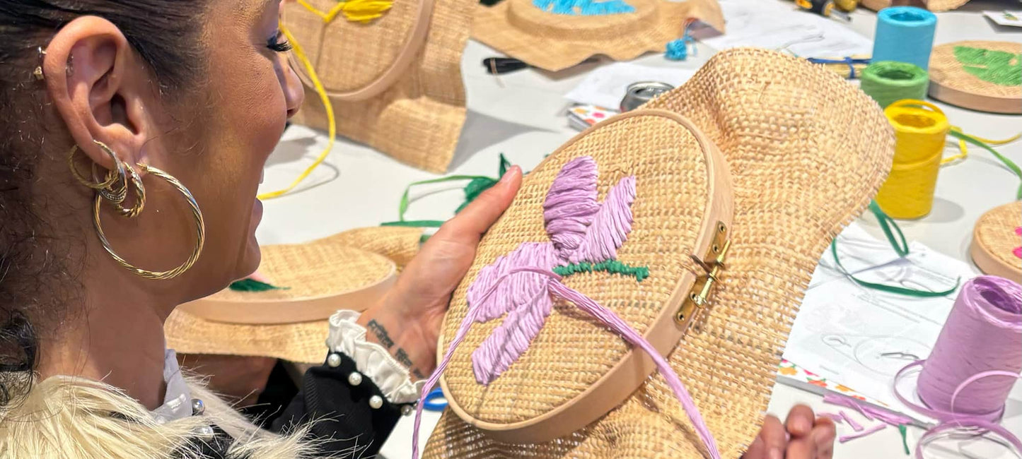 A close-up shot of a woman holding a raffia embroidery hoop with a purple embroidered flower, surrounded by craft supplies and other raffia embroidery hoops on a white table.