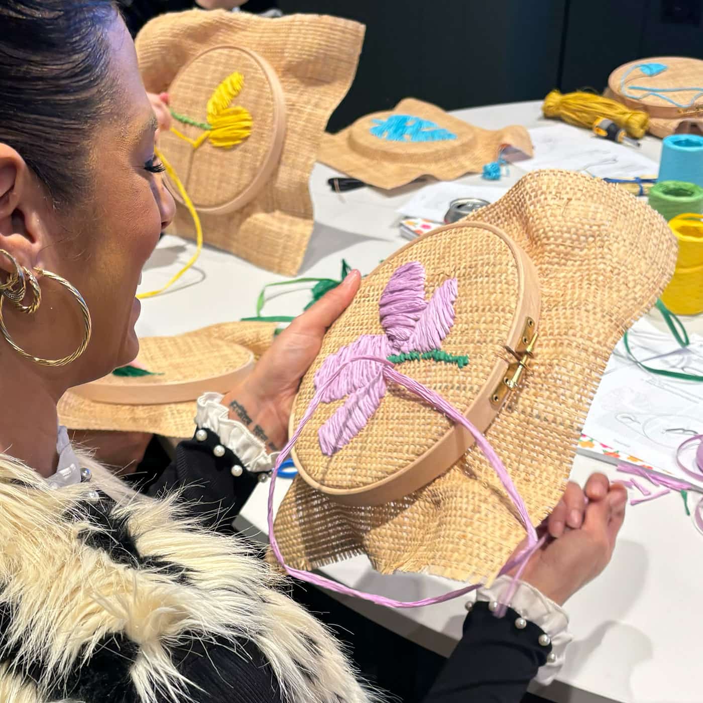 A close-up shot of a woman holding a raffia embroidery hoop with a purple embroidered flower, surrounded by craft supplies and other raffia embroidery hoops on a white table.