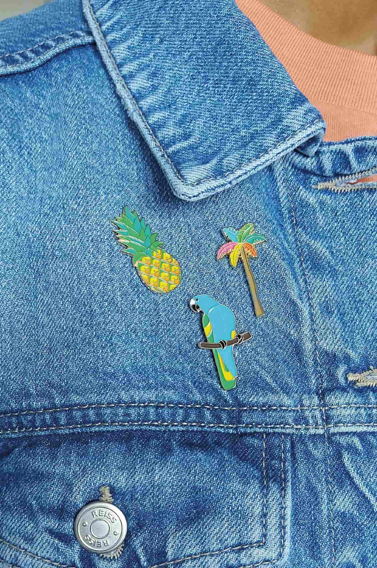 Three colorful enamel pins shaped like a pineapple, a palm tree, and a blue-and-green parrot, displayed on a blue denim jacket over a peach-colored top.