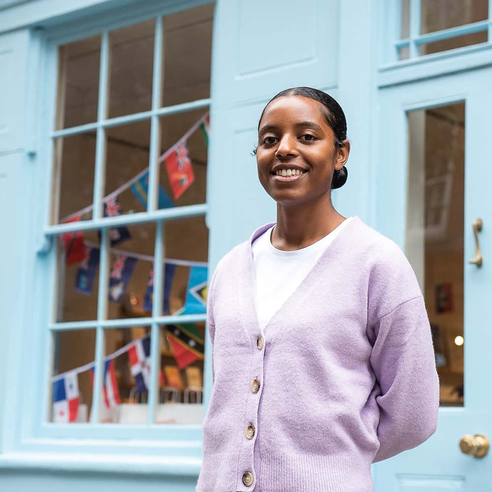 A smiling Tihara Smith with dark hair pulled back stands in front of a light blue building, wearing a lavender cardigan over a white top. The background shows a window with glass panes and Caribbean flags hanging inside.