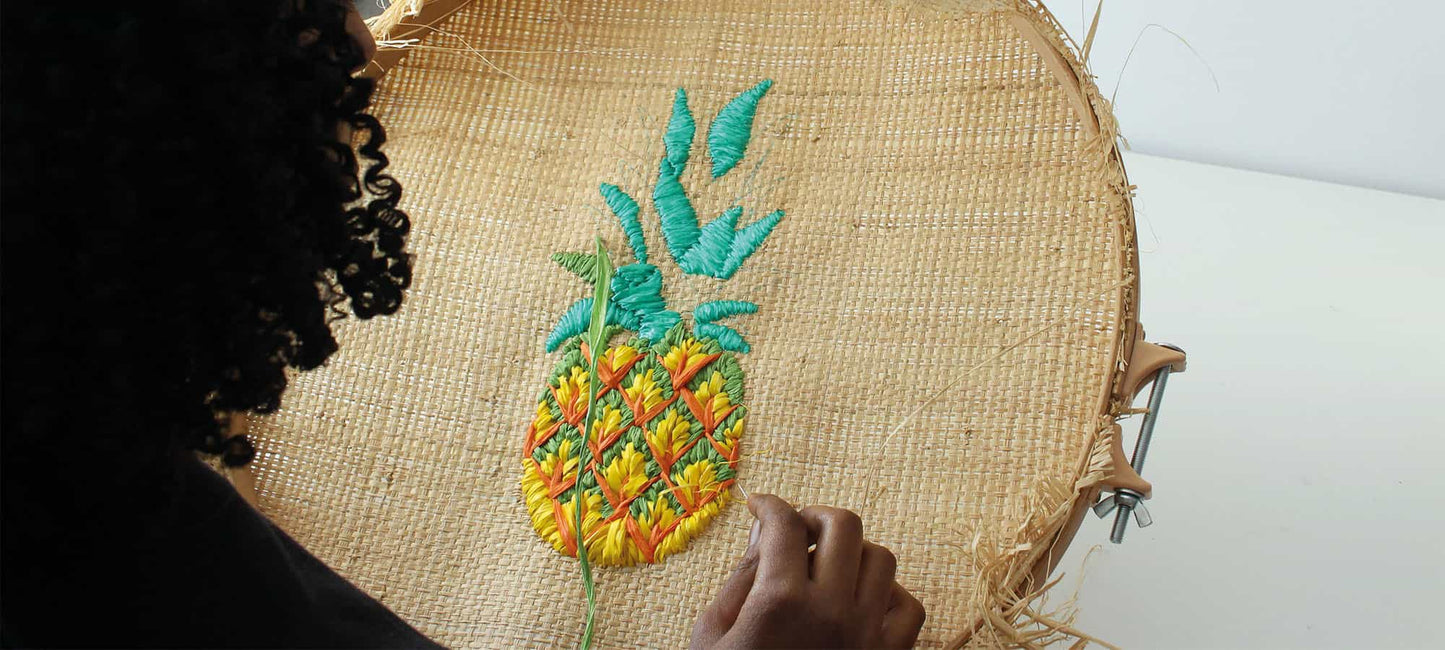 A close-up of a raffia embroidery pineapple design onto a large, circular raffia hoop. Tihara Smith's hand and curly dark hair are visible on the left side, with a green monstera leaf in the background.