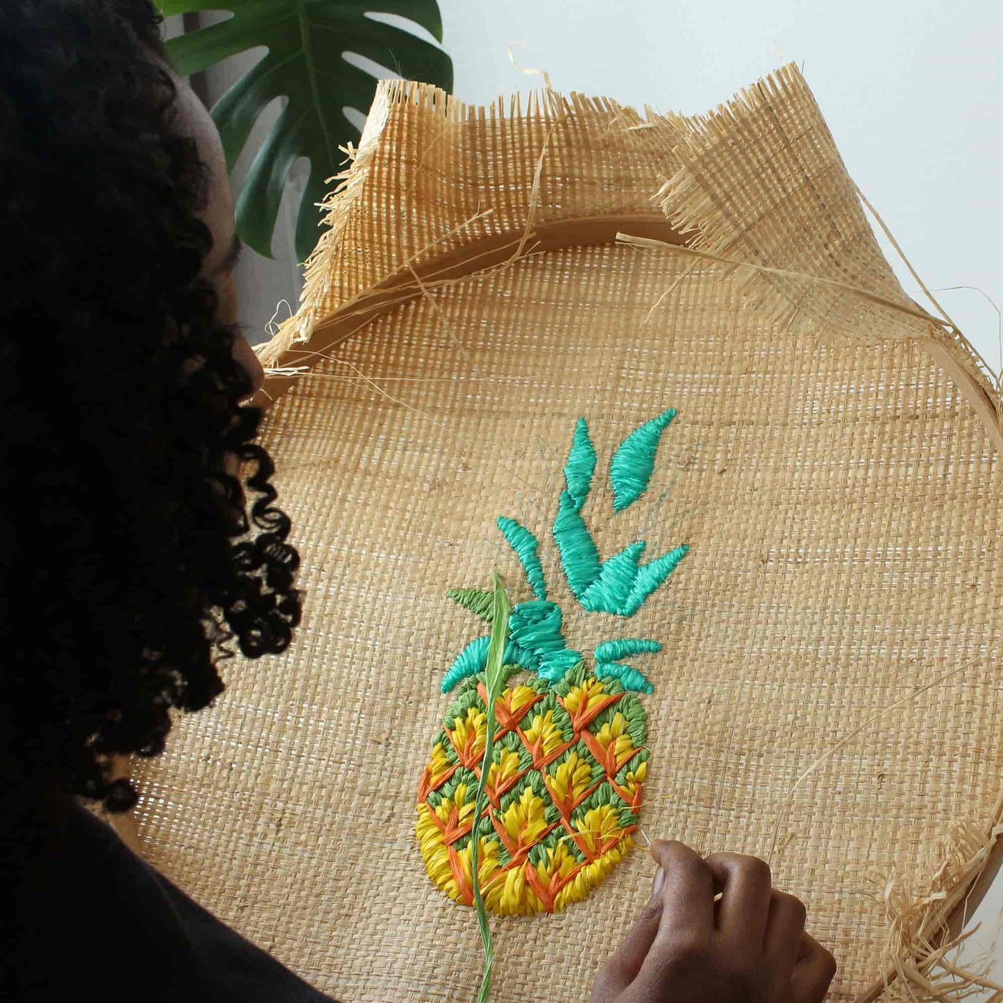 A close-up of a raffia embroidery pineapple design onto a large, circular raffia hoop. Tihara Smith's hand and curly dark hair are visible on the left side, with a green monstera leaf in the background.