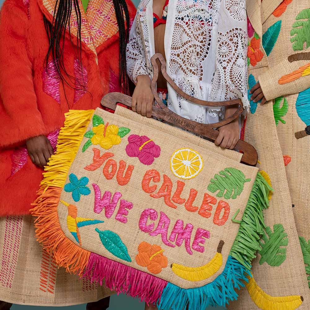 A close-up of a handbag held by two people wearing colourful raffia embroidered clothing. The bag is made of natural raffia with bright multicolour embroidery and fringe, featuring the text 'YOU CALLED WE CAME' and tropical motifs like flowers, leaves, a banana, and a lemon slice.