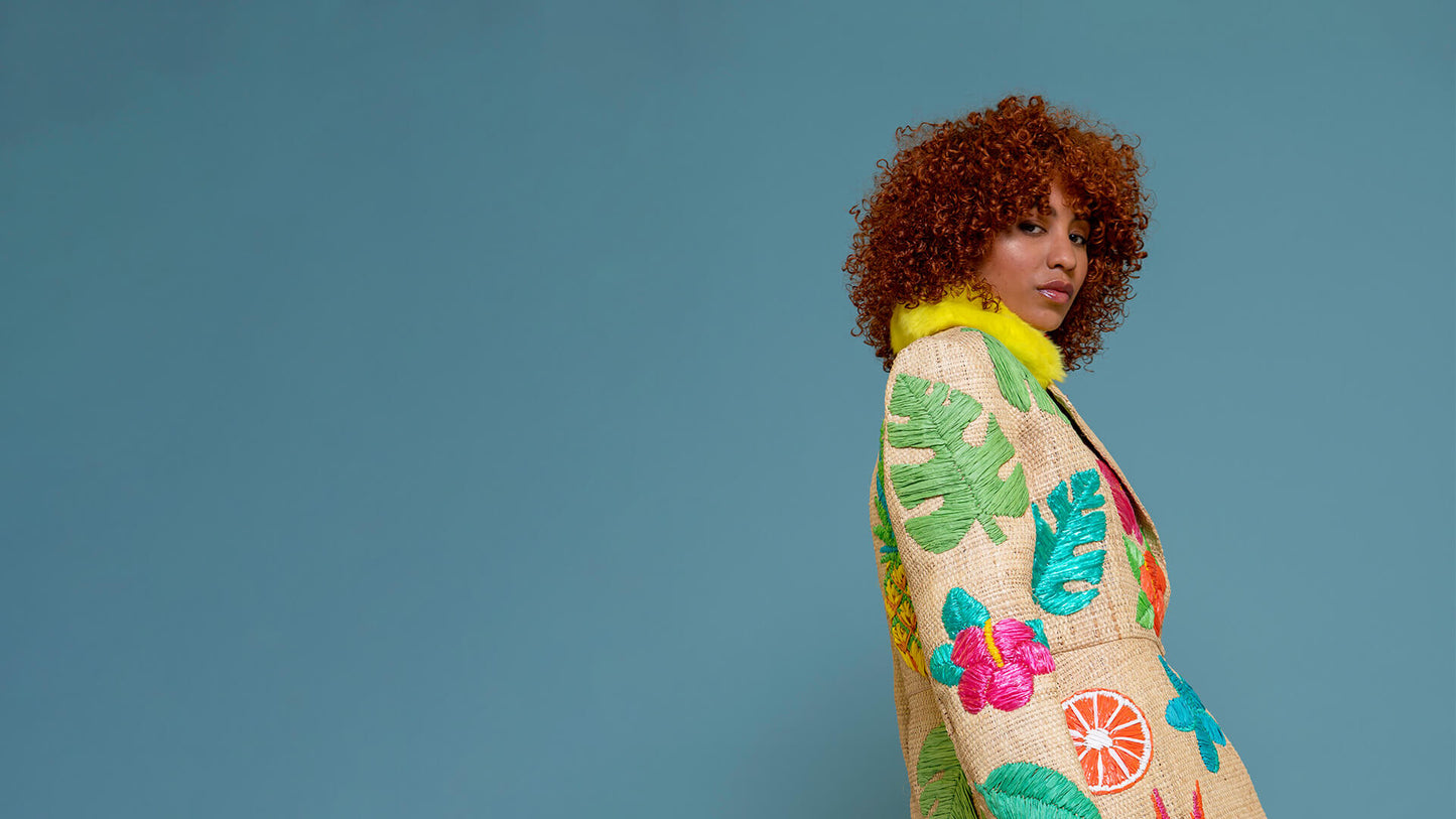 A model with curly red hair is shown from the side, wearing a raffia coat decorated with colourful tropical leaf and floral raffia embroidery, set against a solid blue background.