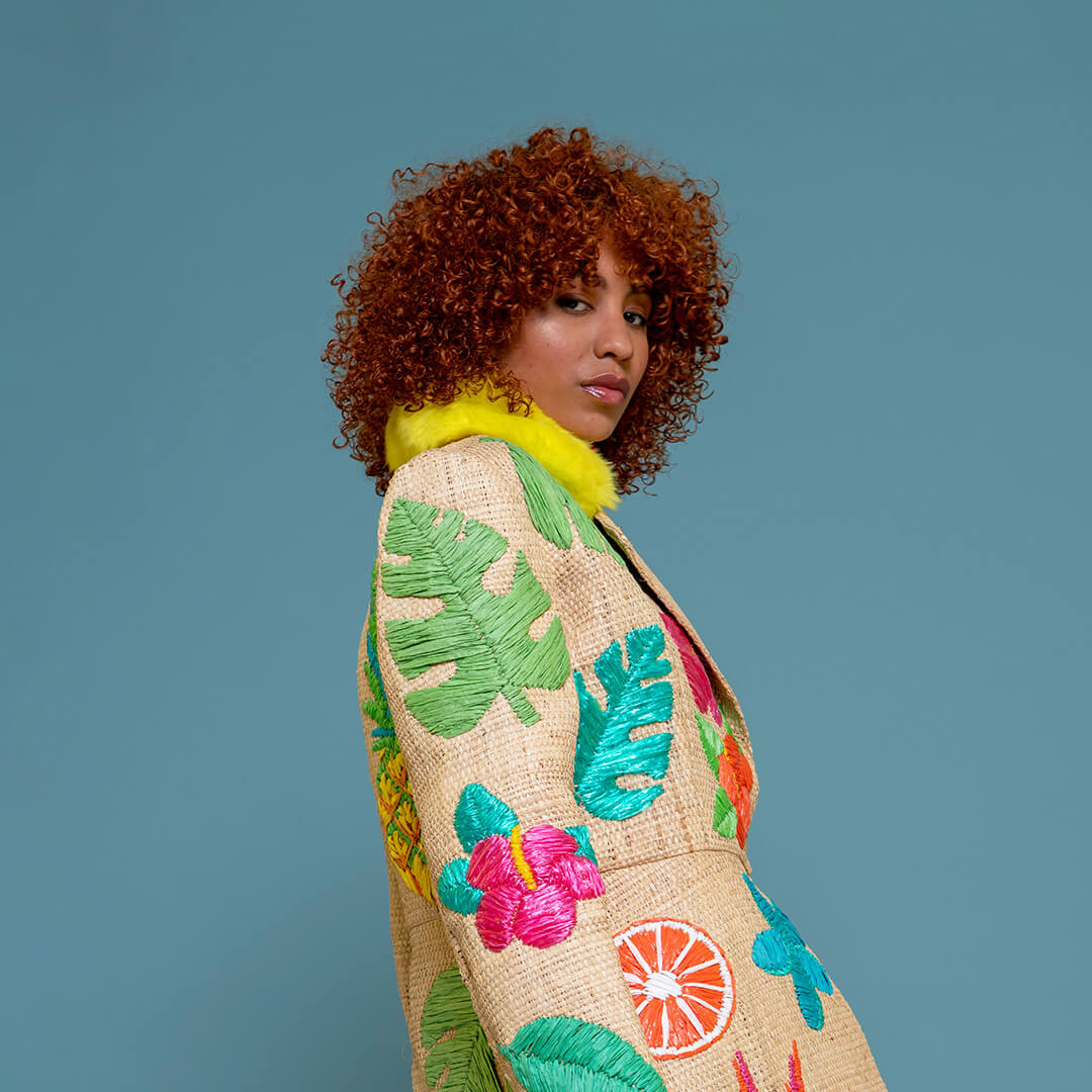 A model with curly red hair is shown from the side, wearing a raffia coat decorated with green tropical leaves, pink flowers, and citrus raffia embroidery motifs, featuring a bright yellow faux-fur collar, set against a solid blue background.