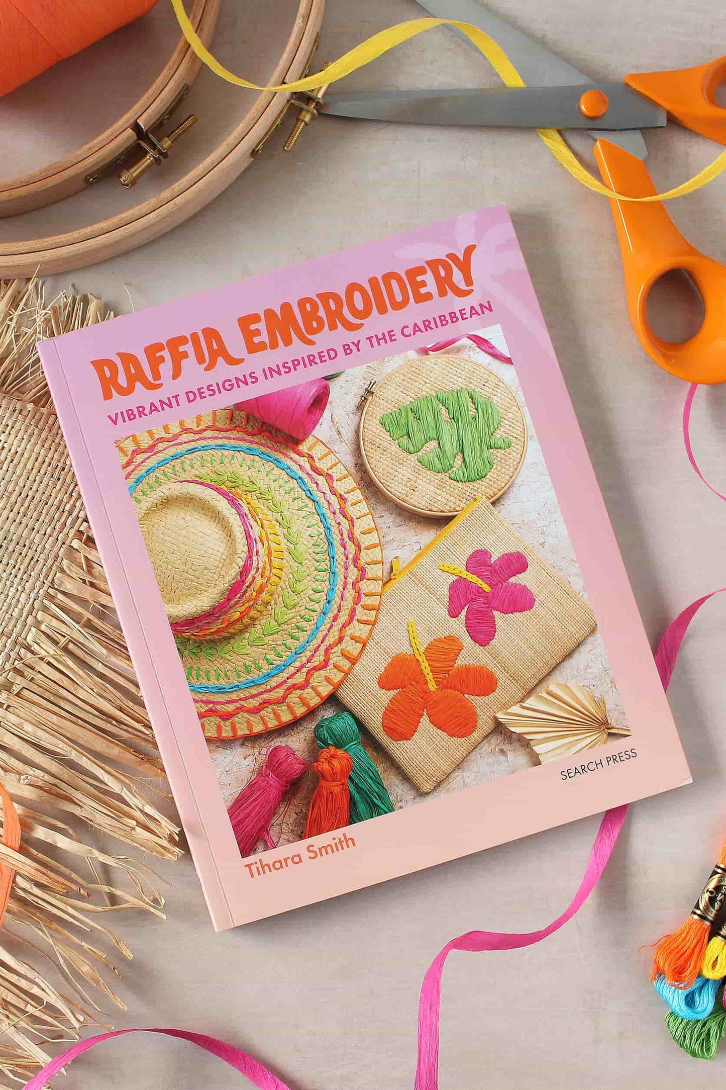 A book titled 'Raffia Embroidery' by Tihara Smith lies on a light surface, surrounded by crafting props including orange-handled scissors, an embroidery hoop, ribbon, and raffia samples. The book cover is pink with colourful raffia embroidery designs shown on the front.