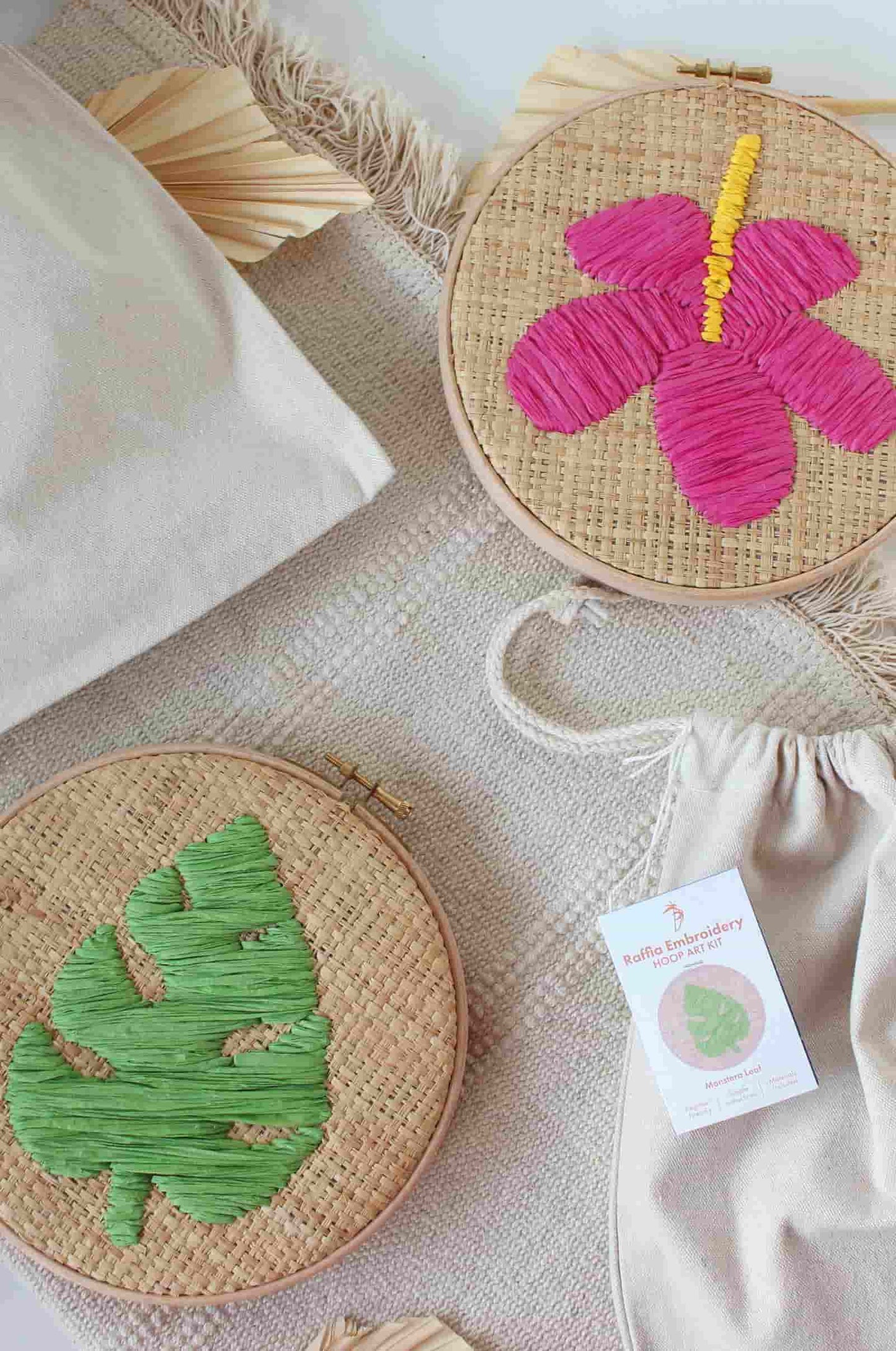 Two round raffia embroidery hoops, one featuring a bright pink flower with a yellow stamen and the other a green leaf, displayed on a textured neutral fabric with a drawstring bag and a product tag.