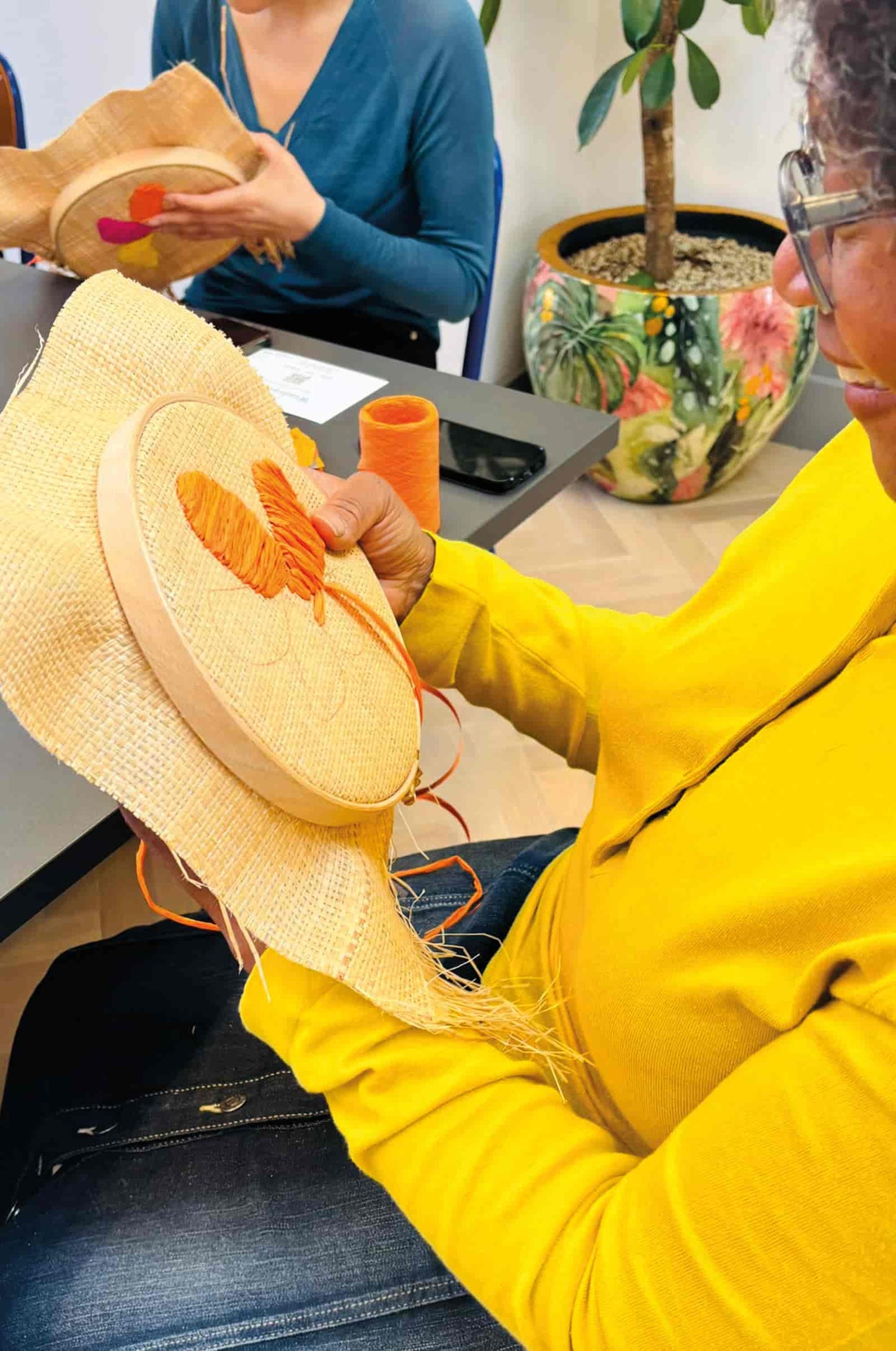 A person in a bright yellow jumper holds a raffia embroidery hoop with orange embroidery inside a wooden hoop, seated at a table in a workshop setting. Another person is visible in the background holding a similar item.